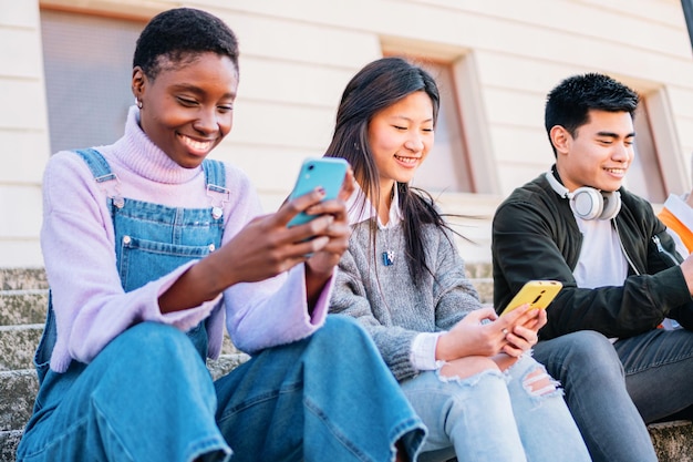 Three multiracial friends chatting with smartphones sitting outdoors and smiling