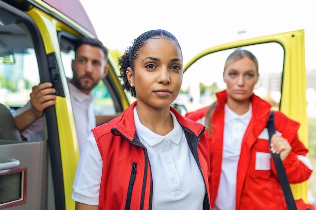 Three multiracial paramedics standing in front of ambulance vehicle carrying portable equipment