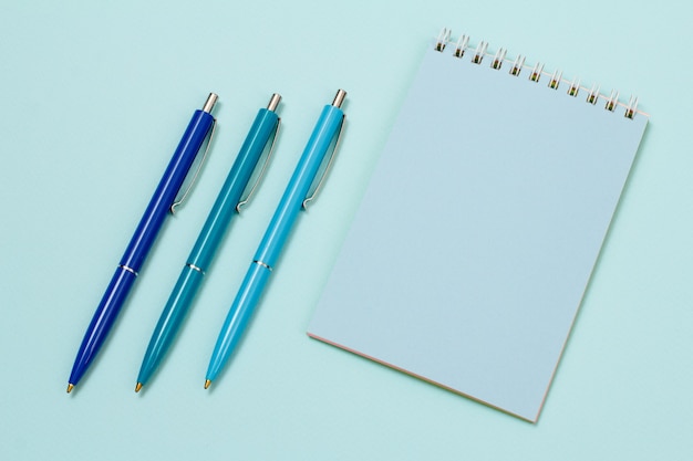 Three pens and notebook on a blue background. Top view.