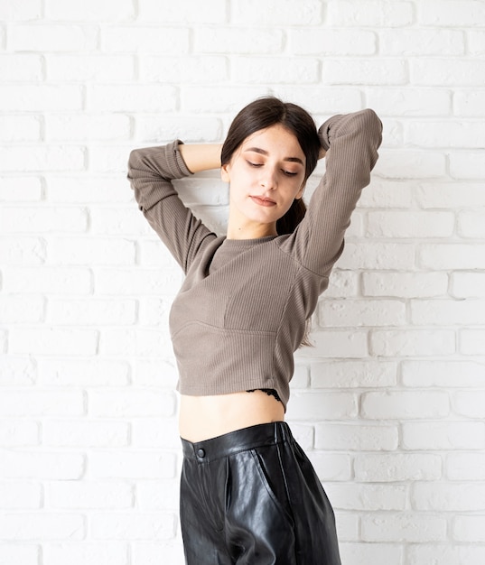 Three quarters length portrait of beautiful smiling brunette woman with long hair wearing brown shirt and black leather shorts, on white brick wall background