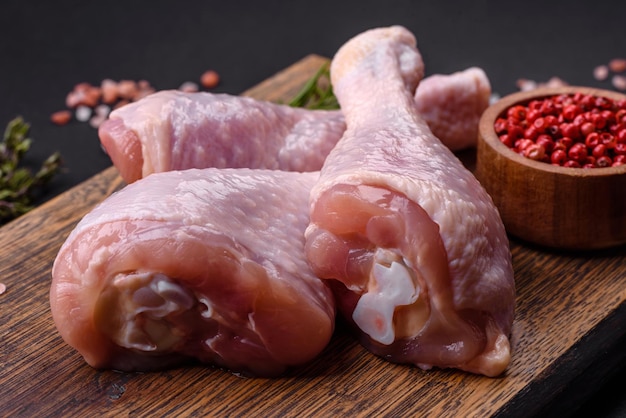 Three raw chicken legs with spices and herbs on a wooden cutting board against a dark concrete background