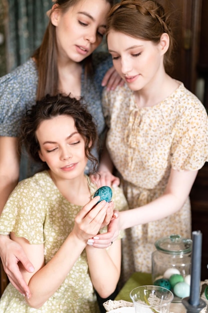 Three sisters beautiful young women ready to celebrate the spring holiday of happy Easter at home decorating dining table with Easter eggs holding in hand vintage countryside fashion style of dress