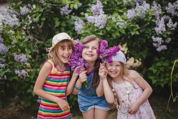 Three smiling european girls stand hugging in a summer garden with lilac flowers in their hands