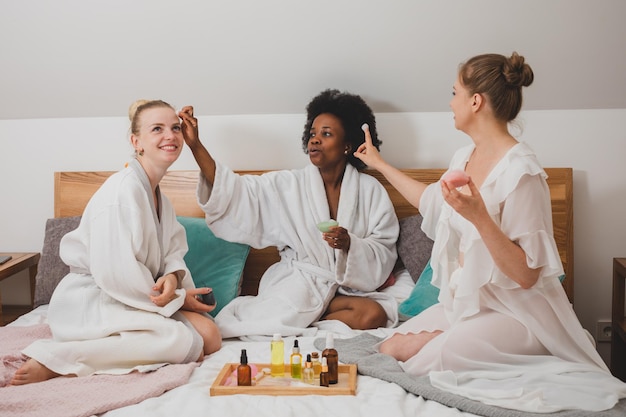 Three smiling girlfriends sit on the bed and smear each other with cream The young women in bathrobes have a spa party