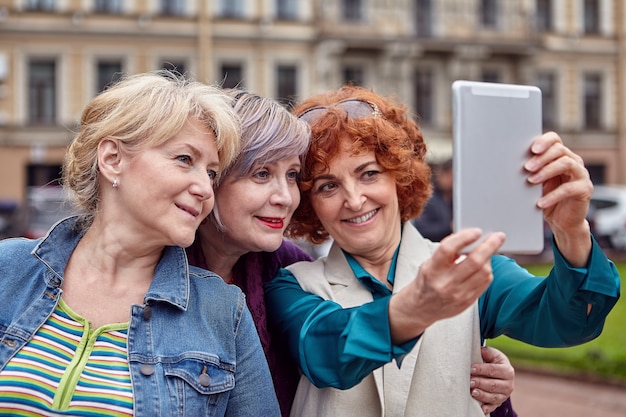 Three smiling older caucasian women make selfie in center of European city with tablet computer.