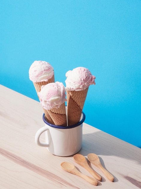 Three strawberry ice cream cones inside a white cup on a blue background with wooden spoons on a wooden table Harsh light with shadows