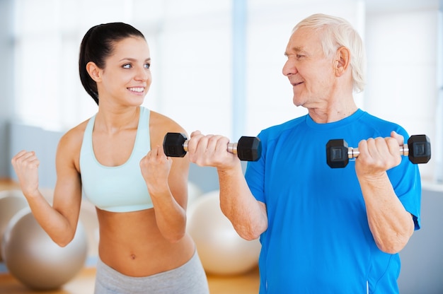 And three times more. Confident female physical therapist showing how to make weight exercise to senior man in health club