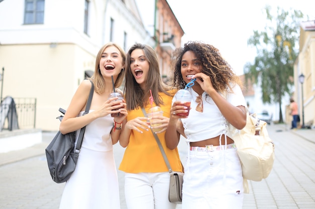 Three trendy cool hipster girls, friends drink cocktail in urban city background.