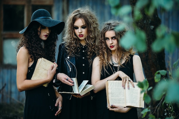 Three vintage women as witches, pose in front of an abandoned building with books in hand on the eve of Halloween