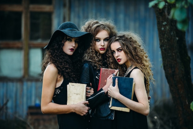 Three vintage women as witches, pose in front of an abandoned building with books in hand on the eve of Halloween