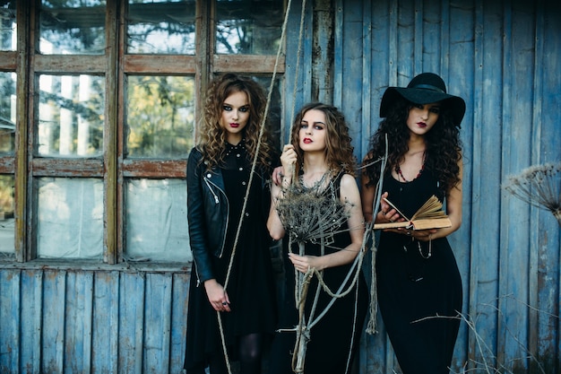 Three vintage women as witches, pose in front of an abandoned building with books in hand on the eve of Halloween