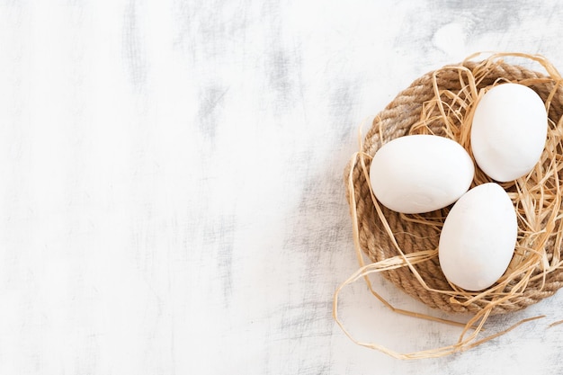 Three white Easter eggs lying on a tray made of twine with some hay White wooden vintage background