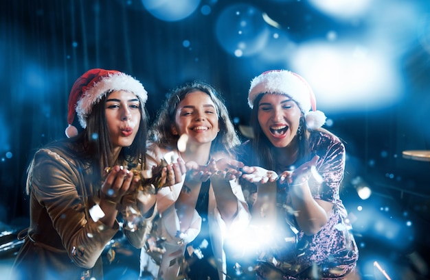 Three women blowing confetti from their hands indoors at holiday time.