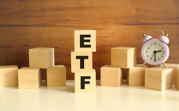 Three wooden cubes stacked vertically on a brown background make up the word ETF. Cubes are scattered nearby and there is a clock. Front View Concepts