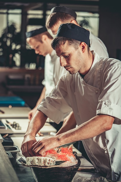 Three young chefs dressed in white uniform decorate ready dish in restaurant they are working on