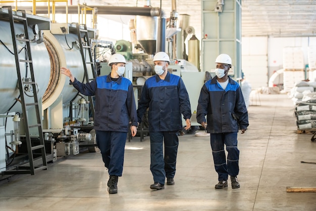 Three young intercultural workers of large contemporary factory moving along industrial plant and discussing new processing equipment