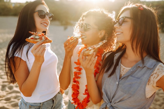 Three young woman spending time together at the beach having picnic eating pizza