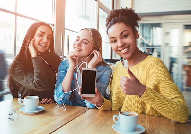 Three young women that are smiling. They closed her eyes and enjoying the music on smartphone. Best friends are sitting in cafe