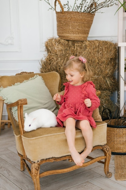 Threeyearold girl in a dress barefoot sits on a chair with a white rabbit in the living room decorated for Easter