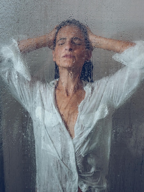 Through glass of charming female model with hand on head showering in transparent shower cabin with wet door during daily routine