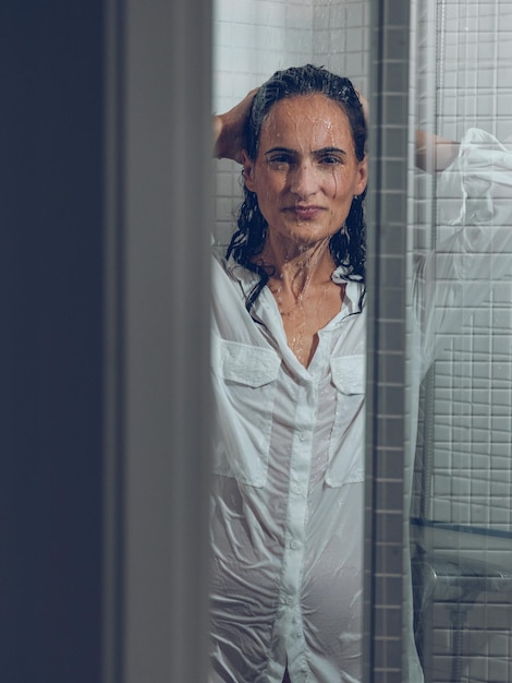 Through glass door attractive alluring female in white wet shirt touching head and looking at camera while standing under waterdrops in shower cabin