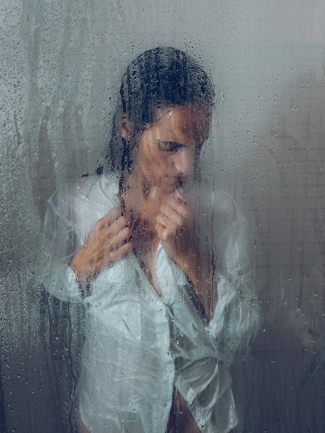 Through glass of sad female in wet blouse looking down in shower cabin near wet transparent door in light bathroom