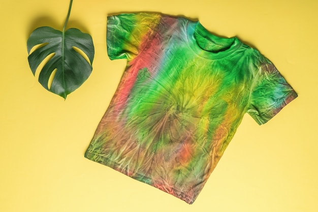 Tie dye style T-shirt on a yellow background with a green leaf. Coloring clothes by hand at home. Flat lay.