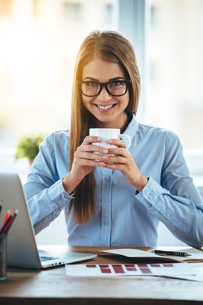 Time for coffee break! Cheerful young beautiful woman holding coffee cup and looking at camera with smile while sitting at her working place