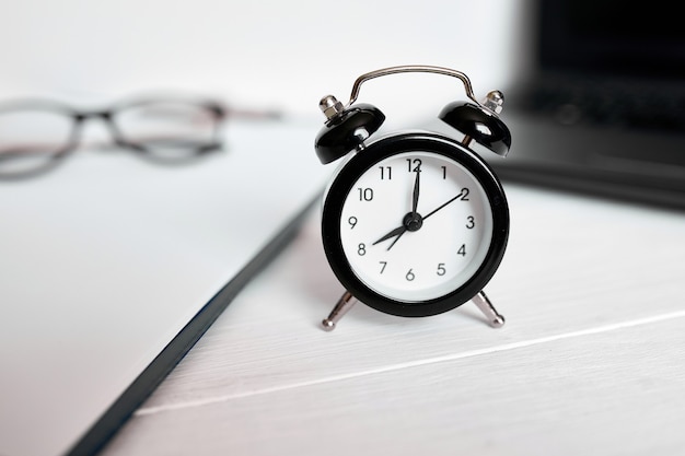 Time management concept, office workplace, notebook, computer, laptop and black alarm clock on white wooden office desk, copy space.