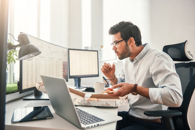 Time for pizza Side view of young hungry trader is eating hot pizza while looking at monitor screen with financial data in his modern office.