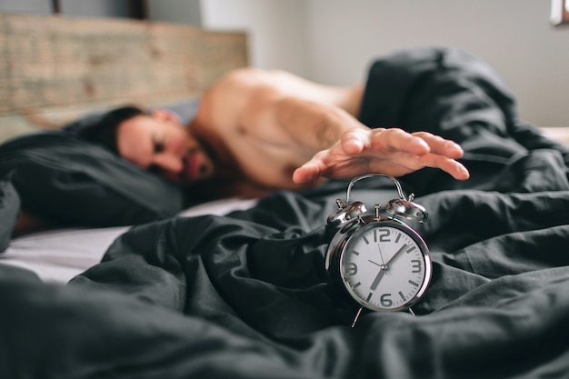 Time to wake up. Tired man in the bed not happy. Mature guy holding alarm clock while checking time for work