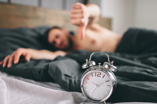 Time to wake up. Tired man in the bed not happy. Mature guy holding alarm clock while checking time for work