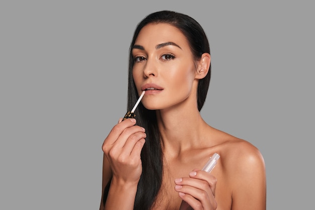 Time for yourself. Attractive young woman applying lip gloss and looking at camera while standing against grey background