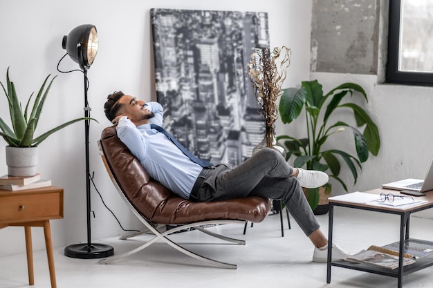 Time for yourself. Business young bearded man with closed eyes smiling vacationer sitting in leather chair sideways to camera