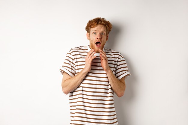 Photo timid young man with red hair, looking scared, jumping and screaming from fear, standing over white background.