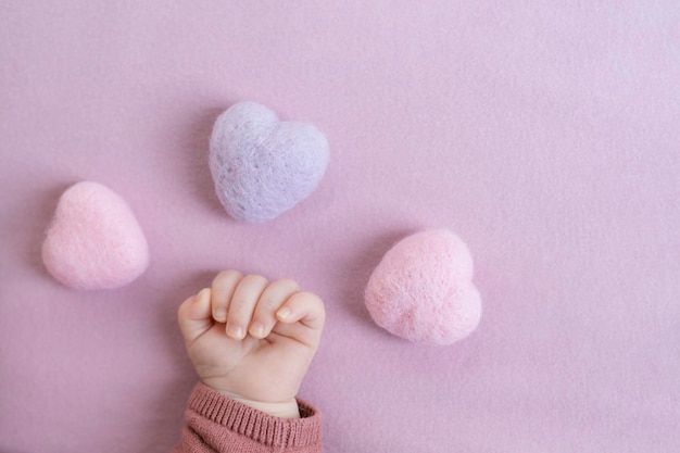 Tiny baby girl hand surrounded by three Heart Shape Wool Felt Balls Bead on light pink background