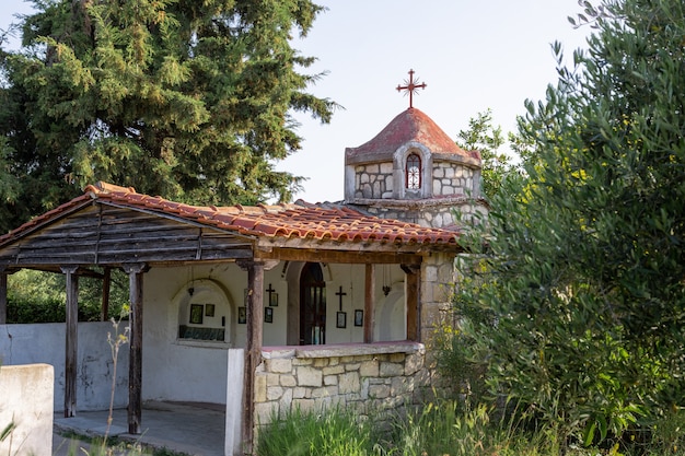 A tiny old orthodox church with stone walls and a tiled roof is surrounded by trees and tall grass