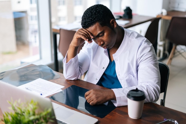 Tired African black male doctor working on laptop thinking about problem and drinking coffee