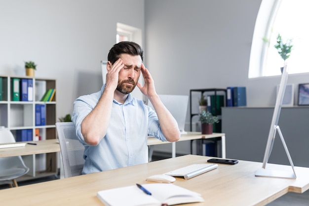 Tired and annoyed young man office worker manager holding his head feeling pain dizziness Sitting at a computer desk in the office