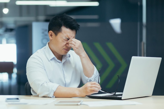 Tired asian businessman working late in office sore eyes man with laptop