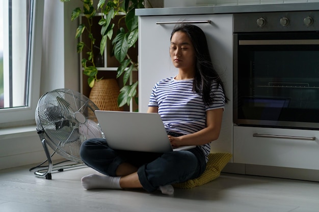 Tired asian girl sits with laptop on floor with air fan suffer from heat at home try to concentrate