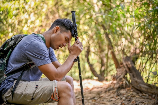 Tired Asian male traveler with trekking gear sits on wooden log resting while trekking