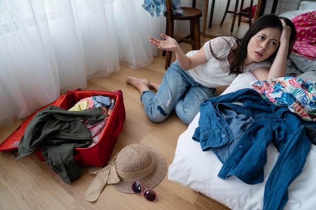 tired asian woman sit on floor near bed with clothes while packing before trip travel concept. angry girl frowning with hand gesturing dislike new year holidays back to home town feeling annoying.