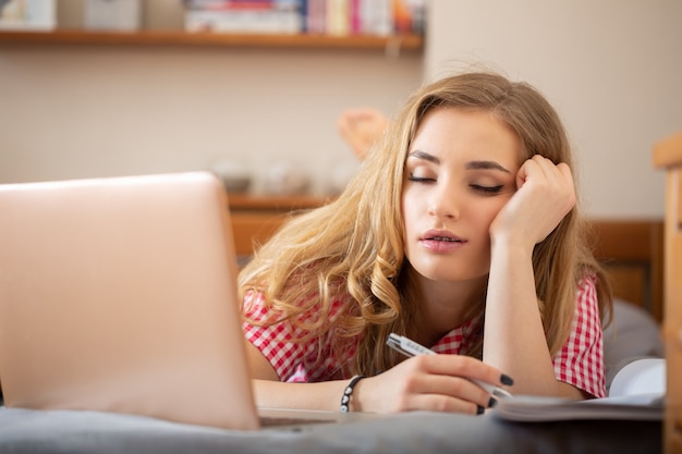 Tired blonde student sleeping during boring online lesson at home due to global covid-19 pandemic.