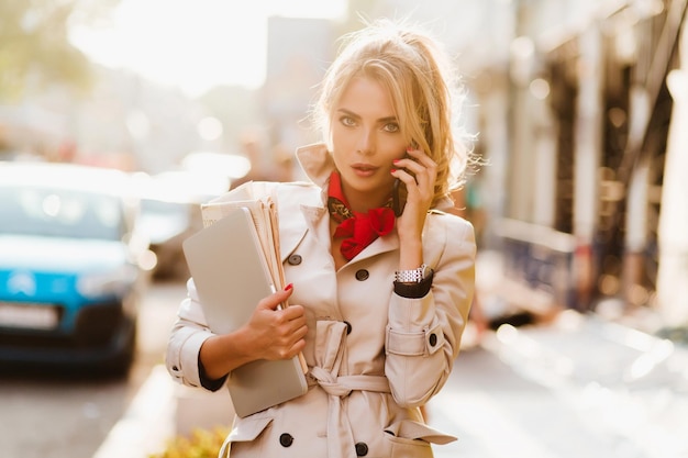 Tired business-lady carrying laptop and fresh newspapers to office in sunny morning. Outdoor portrait of pretty young woman in beige coat talking on phone with serious face expression.