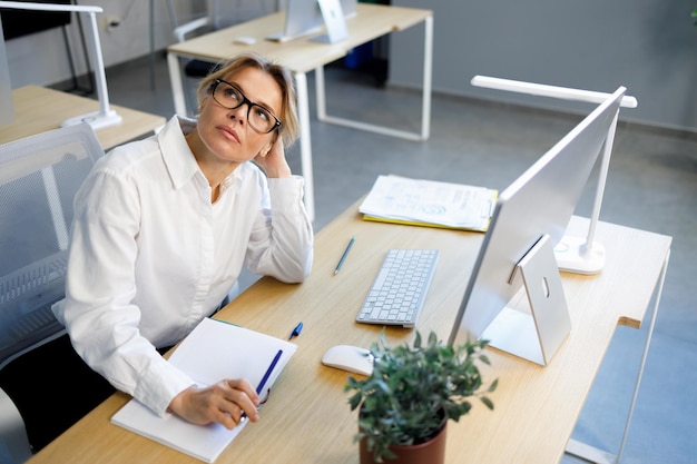 Tired business woman in white shirt and eyeglasses sitting at her desk in the office