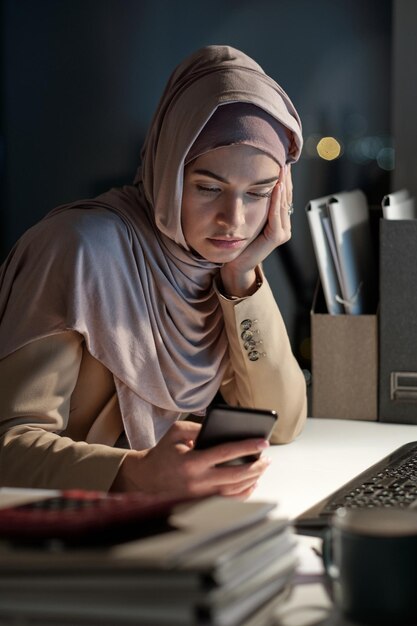 Tired businesswoman in hijab looking through messages in smartphone