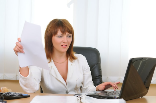 Tired caucasian white businesswoman uses paper sheet as fan and working at laptop in office.