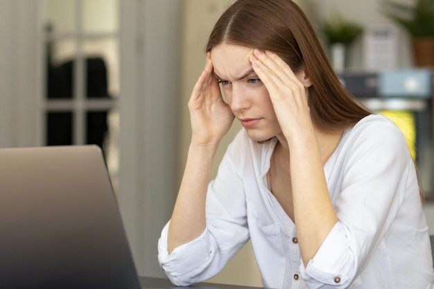 Tired caucasian woman businessman having headache or migraine sitting at the office desk. Young woman freelancer sitting at home office suffering from overwork on computer