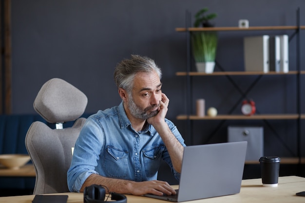 Tired and disappointed senior man working in home office with laptop.